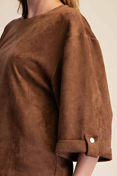 A close-up of a person wearing a brown suede-like top with a rolled cuff and a small white button detail, against a plain light beige background.