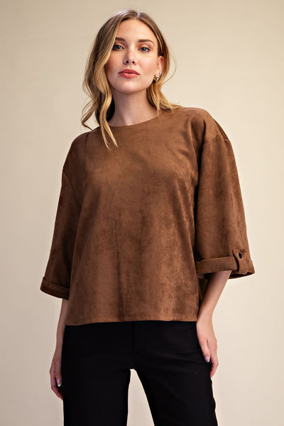 A woman modeling a brown faux-suede cropped top with three-quarter sleeves and rolled cuffs, paired with black pants, against a solid beige background.