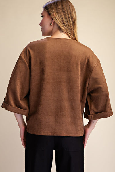 A woman shown from the back wearing a brown suede-like short-sleeve top with rolled cuffs and black pants, against a plain beige studio background.