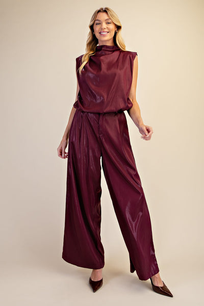 A woman modeling a sleeveless high-neck outfit in a shiny deep burgundy satin-like fabric, photographed against a plain warm beige studio background.