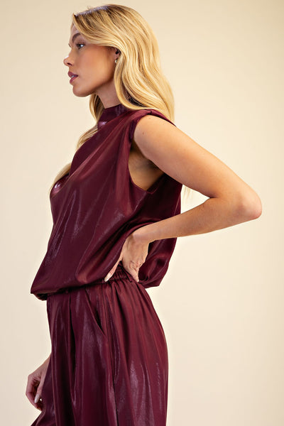 A blonde model wearing a sleeveless, high-neck, dark burgundy outfit, shown from a side profile against a solid beige studio background.