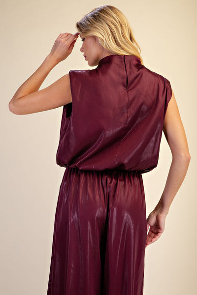 A woman shown from the back wearing a shiny sleeveless burgundy top and matching wide-leg pants against a plain beige studio background.