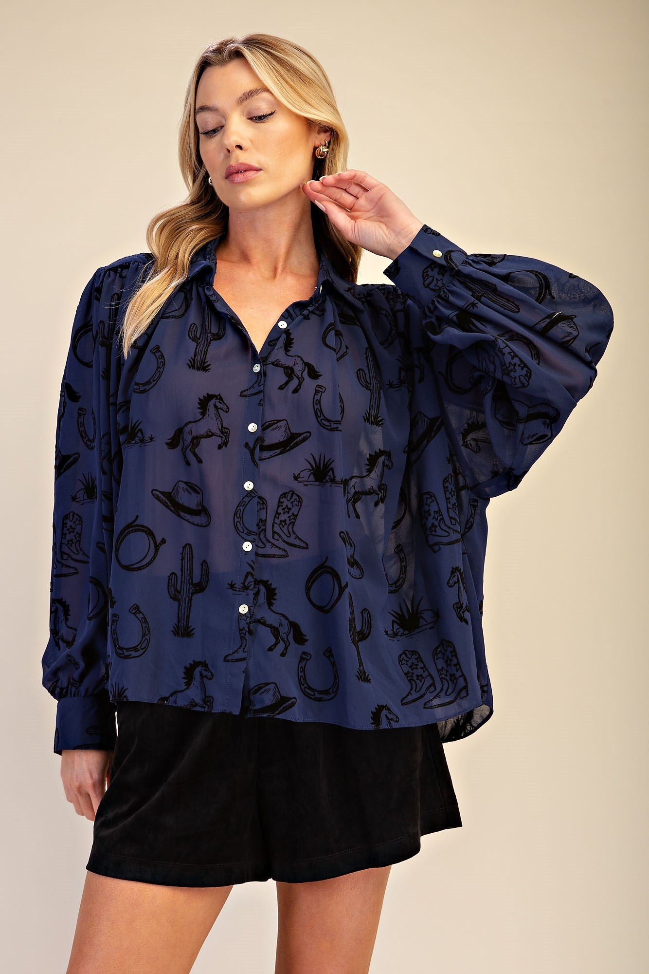 A woman modeling a navy blue long-sleeve button-up blouse with a black western-themed print of cacti, horseshoes, and boots, paired with black shorts against a solid beige background.