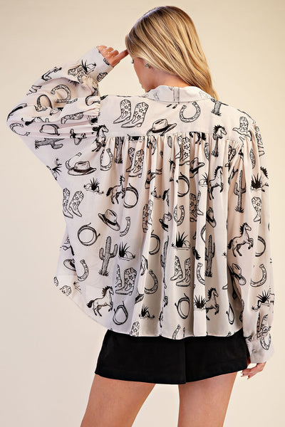 A woman shown from the back wearing a long-sleeve white blouse with a black western-themed print of boots, hats, horseshoes, and cacti, paired with black shorts, against a solid beige background.