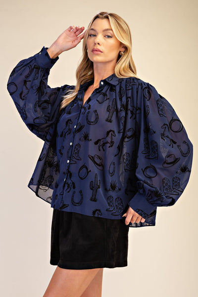 A woman modeling a sheer navy blue button-up blouse with a black western-themed print of horseshoes, boots, cacti, and saddlery, paired with a black skirt against a solid beige background.