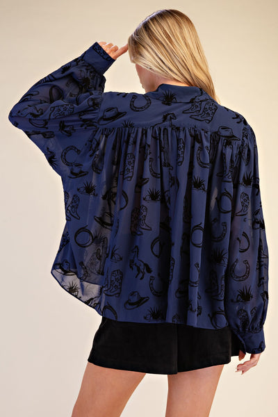 A woman shown from the back wearing a navy blue long-sleeve blouse with a black cowboy-themed pattern (including boots, hats, horseshoes, and cactus silhouettes) and black shorts, set against a plain beige studio background.