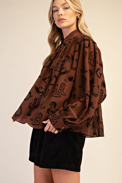 A woman wearing a brown long-sleeve blouse with a black western-themed print of boots, hats, and cacti, paired with black shorts, against a solid beige background.