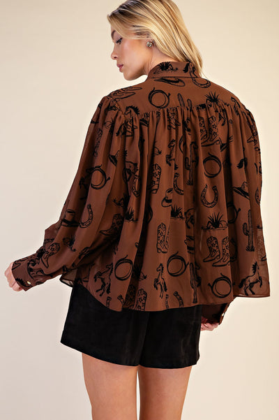 A woman shown from the back wearing a brown long-sleeve blouse with a black western-themed print of horseshoes, cacti, and boots, paired with black shorts, against a solid beige background.