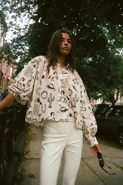 A woman standing on a city sidewalk wearing a cream-colored long-sleeve blouse with a black western-themed print of cacti, boots, and hats, paired with white pants.