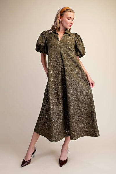 A woman models a mid-length olive green dress with a textured, tonal leaf-like pattern, featuring short puff sleeves and a collared neckline. She is wearing dark brown pointed-toe heels against a solid light beige studio background.