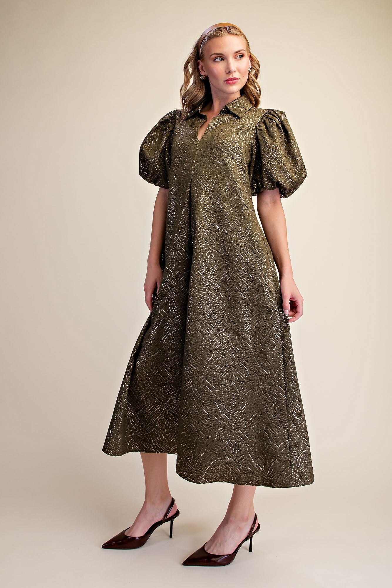 A woman modeling a midi-length olive green dress with a textured, metallic-looking pattern, puffed short sleeves, and a split neckline. She is wearing dark brown slingback heels against a solid beige studio background.