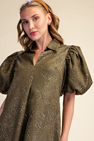 A woman wearing a metallic olive green short-sleeve blouse with a textured jacquard pattern, puffed bubble sleeves, and a split V-neckline, set against a plain beige background.
