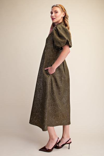A woman modeling a midi-length olive green dress with a textured, embossed pattern, puffed short sleeves, and side pockets, paired with dark red slingback heels against a solid beige background.