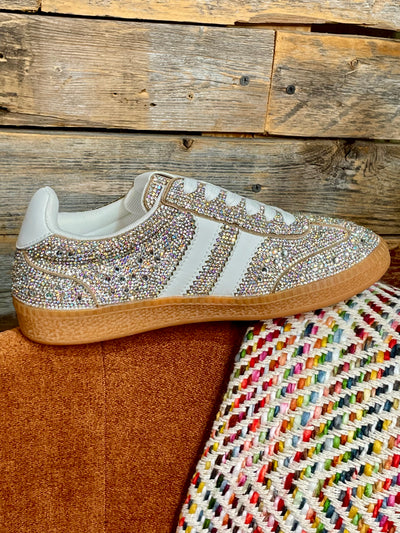 A low-top sneaker covered in sparkling rhinestones, featuring white side stripes, a white heel tab, and a brown gum sole, resting on a brown suede cushion next to a colorful woven textile against a rustic wood plank background.