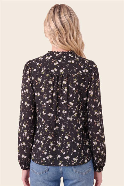 A woman viewed from the back wearing a long-sleeve black blouse with a dense white and light green floral print, paired with blue jeans, against a solid light beige background.
