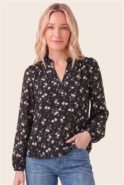 A woman wearing a black long-sleeve blouse with a small white and yellow floral print, paired with blue jeans, against a solid light beige background.