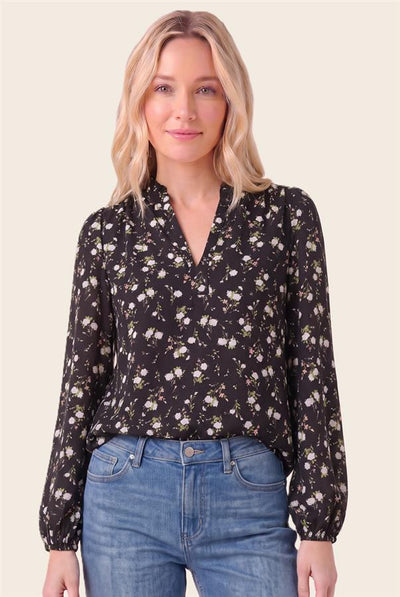A woman modeling a black long-sleeve blouse with a white and yellow floral print, paired with blue jeans, against a solid light beige background.