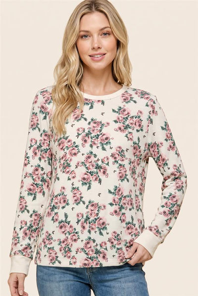A woman modeling a long-sleeve crewneck top with a pink and green floral print on a cream background, paired with blue jeans against a solid beige studio background.