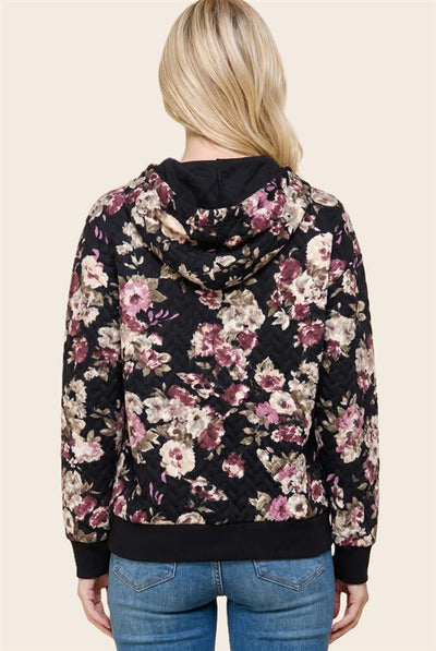 A woman viewed from the back wearing a black floral hooded sweatshirt with pink, purple, and beige flowers, paired with blue jeans, against a solid light beige background.