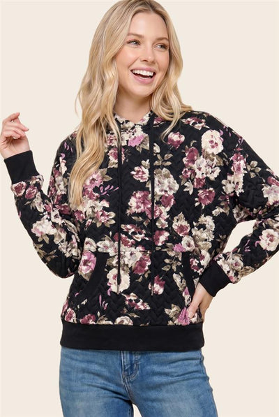 A woman wearing a black floral print hoodie with pink, purple, and beige flowers, paired with blue jeans, against a solid light beige background.