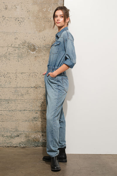 Woman wearing a denim jumpsuit standing against a textured wall.