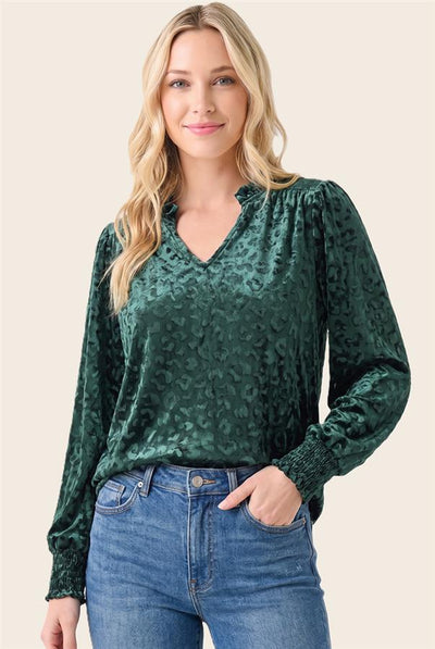 A woman wearing a dark green leopard-print long-sleeve blouse with a V-neckline, paired with blue jeans, against a solid light beige background.