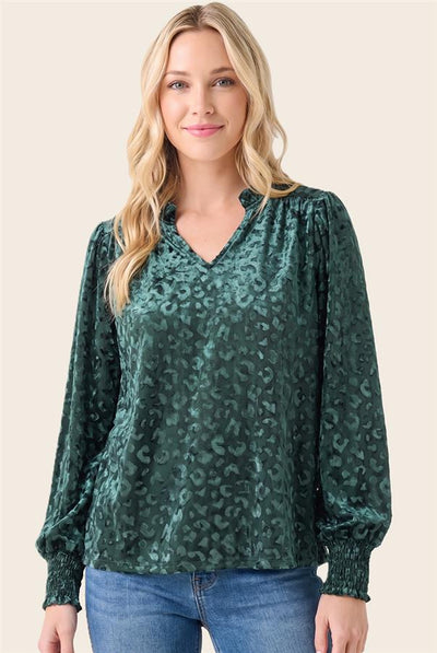 A woman wearing a dark green leopard-print long-sleeve blouse with a V-neckline, paired with blue jeans, against a solid light beige background.