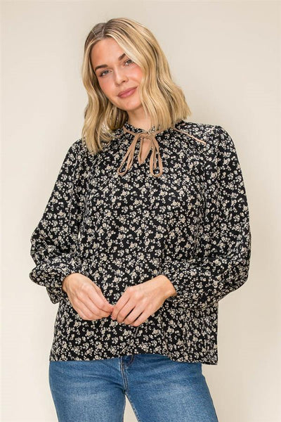 A woman wearing a black long-sleeve blouse with a white floral print and tie-neck detail, paired with blue jeans, standing against a plain beige background.