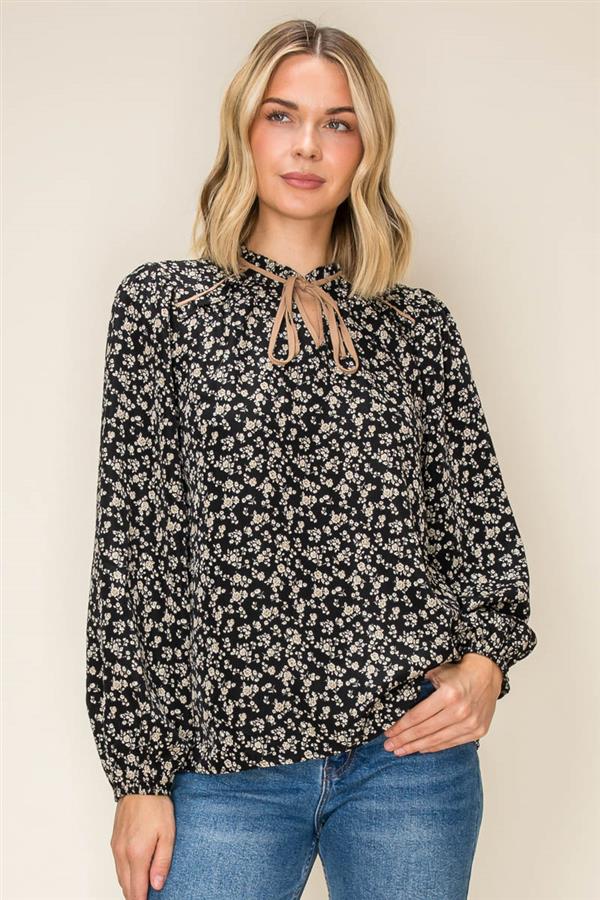A woman wearing a black long-sleeve blouse with a dense white floral print and a tie-neck detail, paired with blue jeans, against a plain beige background.