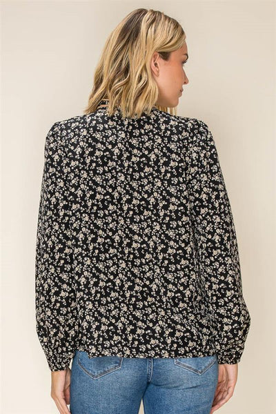 A woman shown from the back wearing a black long-sleeve blouse with a dense small floral print in light beige or off-white, paired with blue jeans, against a plain beige studio background.