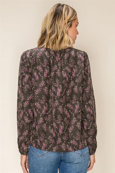 A woman viewed from the back wearing a dark olive long-sleeve blouse with a pink floral pattern and blue jeans, set against a plain beige background.