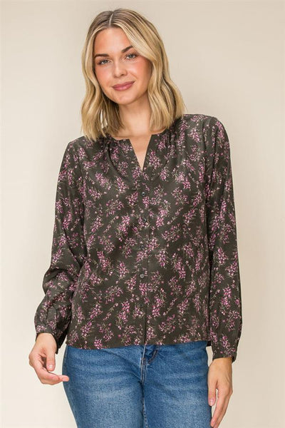 A woman wearing a dark olive long-sleeve blouse with a pink floral pattern and blue jeans, standing against a plain beige background.