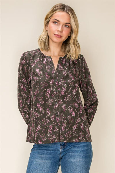 A woman wearing a dark olive long-sleeve blouse with a pink floral pattern and a split neckline, paired with blue jeans, against a plain beige background.