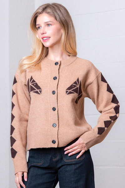 Woman wearing a beige cardigan with brown patterns against a white brick wall.