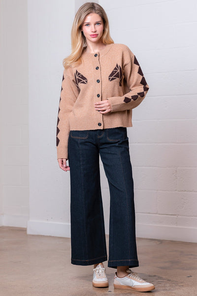 Woman wearing a beige cardigan with black geometric patterns and dark blue jeans against a white wall.