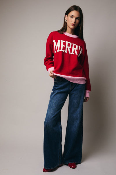 Woman wearing a red 'MERRY' sweater and blue jeans against a plain background