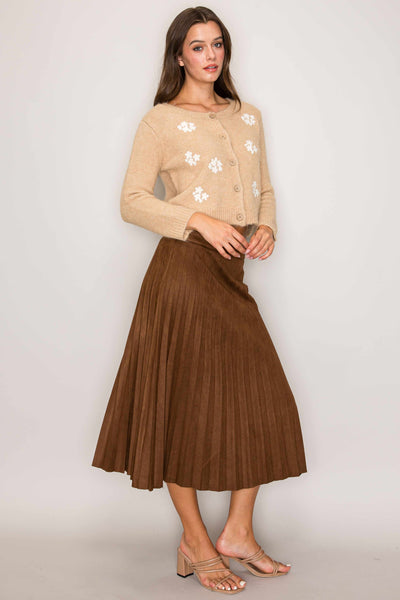 Woman wearing a beige cardigan with floral patterns and a brown pleated skirt on a plain background