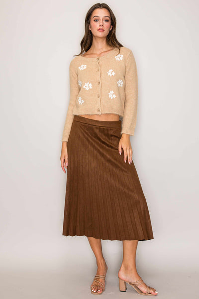 Woman wearing a beige cardigan with floral patterns and a brown pleated skirt on a plain background