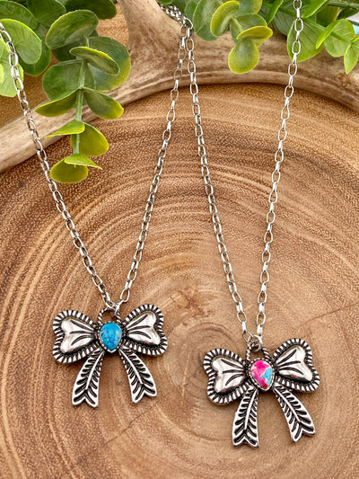 Two bow necklaces on a wooden surface with greenery. One is pink and one is blue.