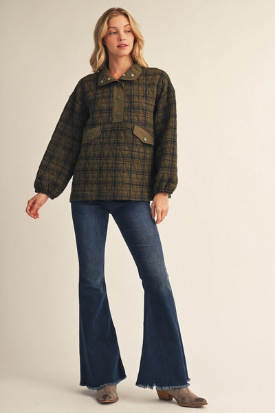 A woman modeling a dark olive green and black plaid button-up jacket with front flap pockets, paired with dark blue flared jeans and brown boots, against a solid light beige background.