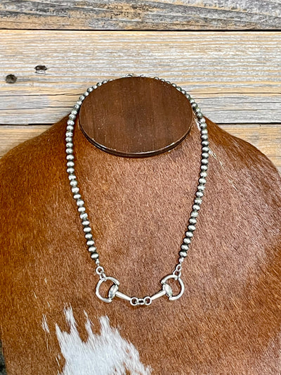 Silver beaded necklace with a horse bit on a brown and white cowhide surface.