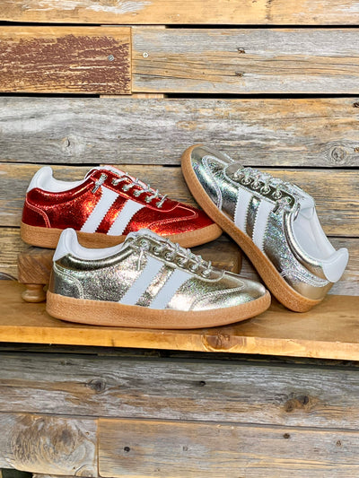 Three low-top sneakers displayed on a rustic wooden shelf: one metallic red, one metallic silver, and one metallic gold, all with white side stripes, set against a weathered wood plank background.