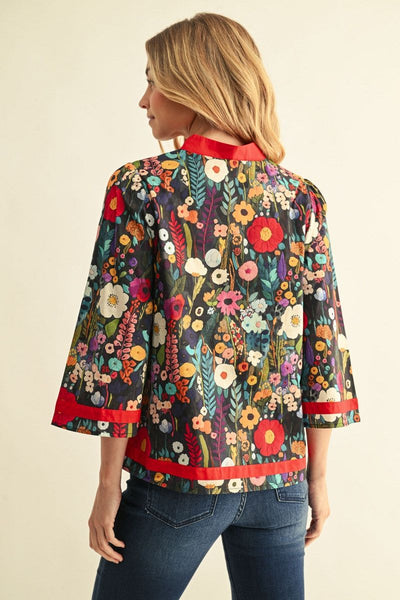 A woman shown from the back wearing a short-sleeve floral jacket with red trim, paired with dark blue jeans, against a plain beige background.