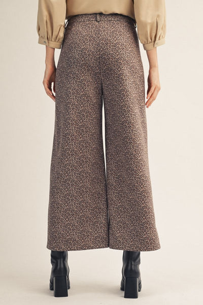 Back of a person wearing high-waisted, wide-leg cropped trousers with an allover brown and beige animal print, styled with a beige top and black ankle boots against a plain beige studio background.