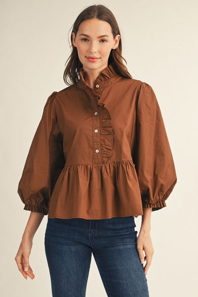 A woman modeling a brown long-sleeve blouse with a high ruffled collar, button front, and gathered peplum waist, paired with blue jeans against a plain light beige studio background.
