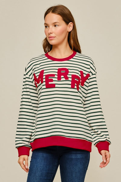 A woman wearing a festive long-sleeve crewneck sweater with green and white horizontal stripes, red ribbed trim, and 'MERRY' written across the chest in red letters, paired with blue jeans against a solid beige background.
