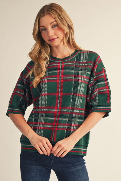A woman wearing a green plaid short-sleeve knit top with red and white stripes, paired with dark blue jeans, against a solid beige background.
