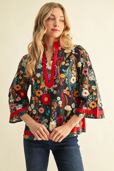 A woman wearing a bohemian-style floral blouse with red trim, paired with dark blue jeans, standing against a plain beige background.
