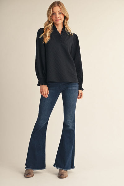 A woman modeling a black long-sleeve textured blouse with a ruffled collar, paired with dark blue flare jeans and brown shoes, against a solid light beige background.