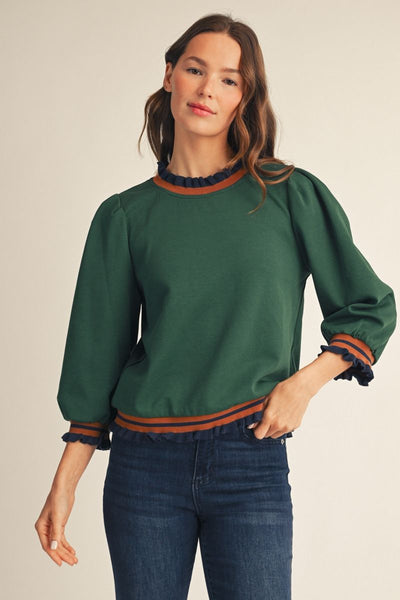 A woman wearing a dark green long-sleeve top with brown and navy ruffled trim at the collar, cuffs, and hem, paired with dark blue jeans, against a plain beige background.